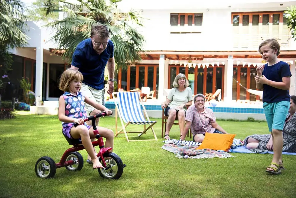 A joyful family enjoys a sunny afternoon in their lush, well-maintained backyard; a young girl rides a red tricycle with help from her grandfather, while others relax on a picnic blanket and deck chairs, surrounded by neatly trimmed grass—showcasing the results of professional lawn maintenance services.