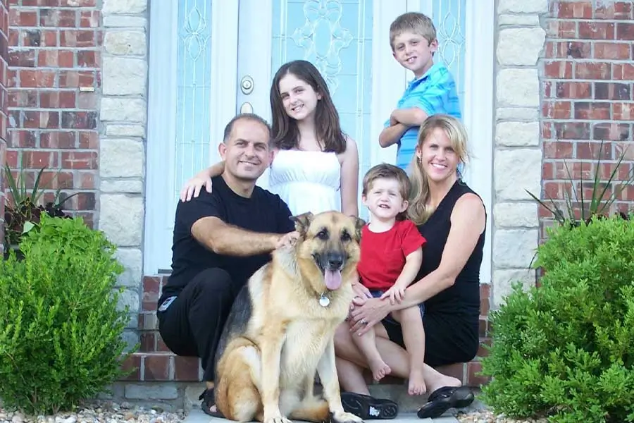 Family of five with their German Shepherd dog, smiling and posing in front of their brick house, enjoying a beautifully maintained lawn. Learn more about Green T Lawn Care and their expert services.