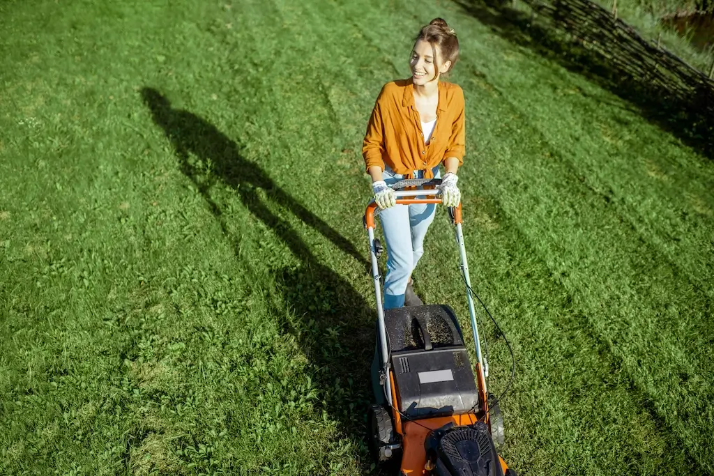 A woman smiling while cutting grass with an electric lawn mower. Regular mowing is an essential part of how to care for lawn in fall, as it helps maintain grass health before the season changes.