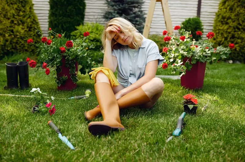 A woman in orange gardening boots sits on a well-trimmed, green lawn, surrounded by potted red flowers and gardening tools. She looks tired but content after working in her backyard, which showcases the lush results of consistent Gaithersburg MD lawn care. The healthy grass and vibrant plants create a picture of suburban gardening success.