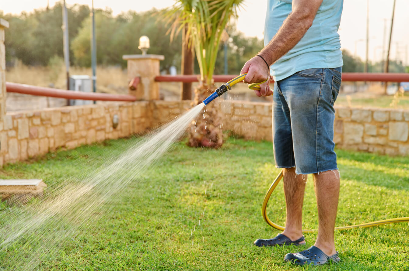 A man in casual summer clothes watering a green lawn with a yellow garden hose, demonstrating a foundational step in maintaining healthy grass that is an important practice in lawn care tips for beginners.
