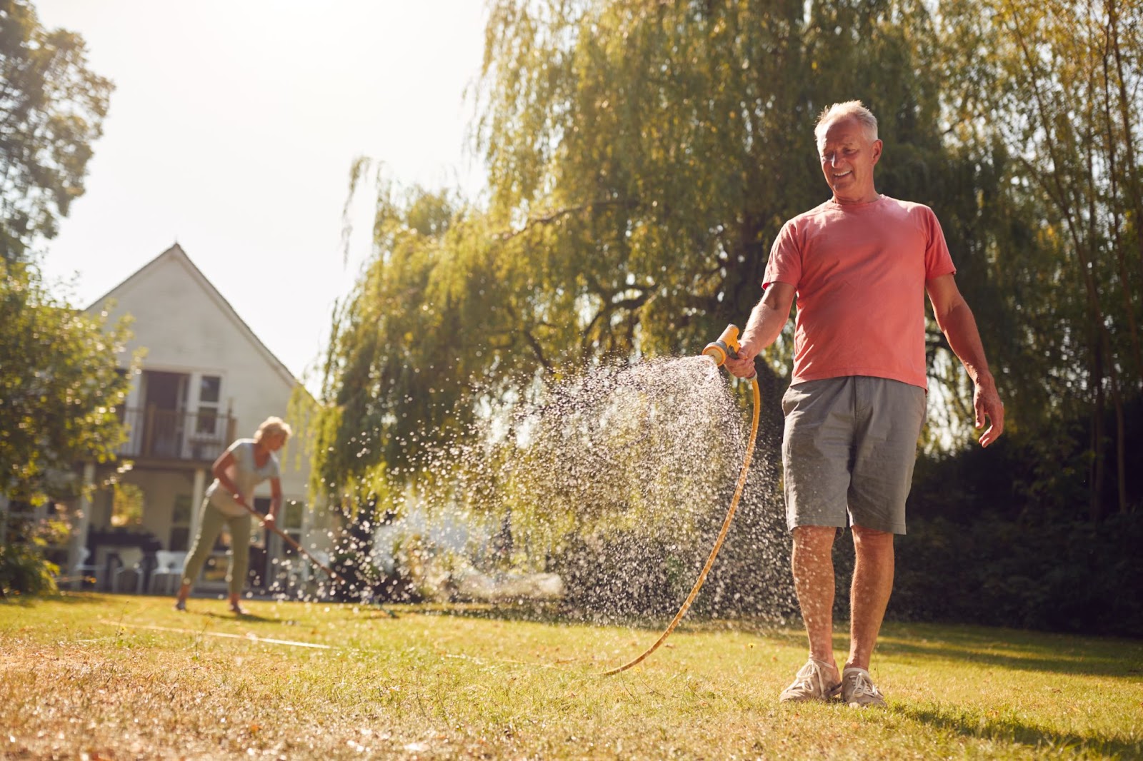 Smiling man in casual summer clothing watering his backyard lawn with a hose on a sunny day, illustrating part of a summer lawn treatment routine for healthy grass maintenance.