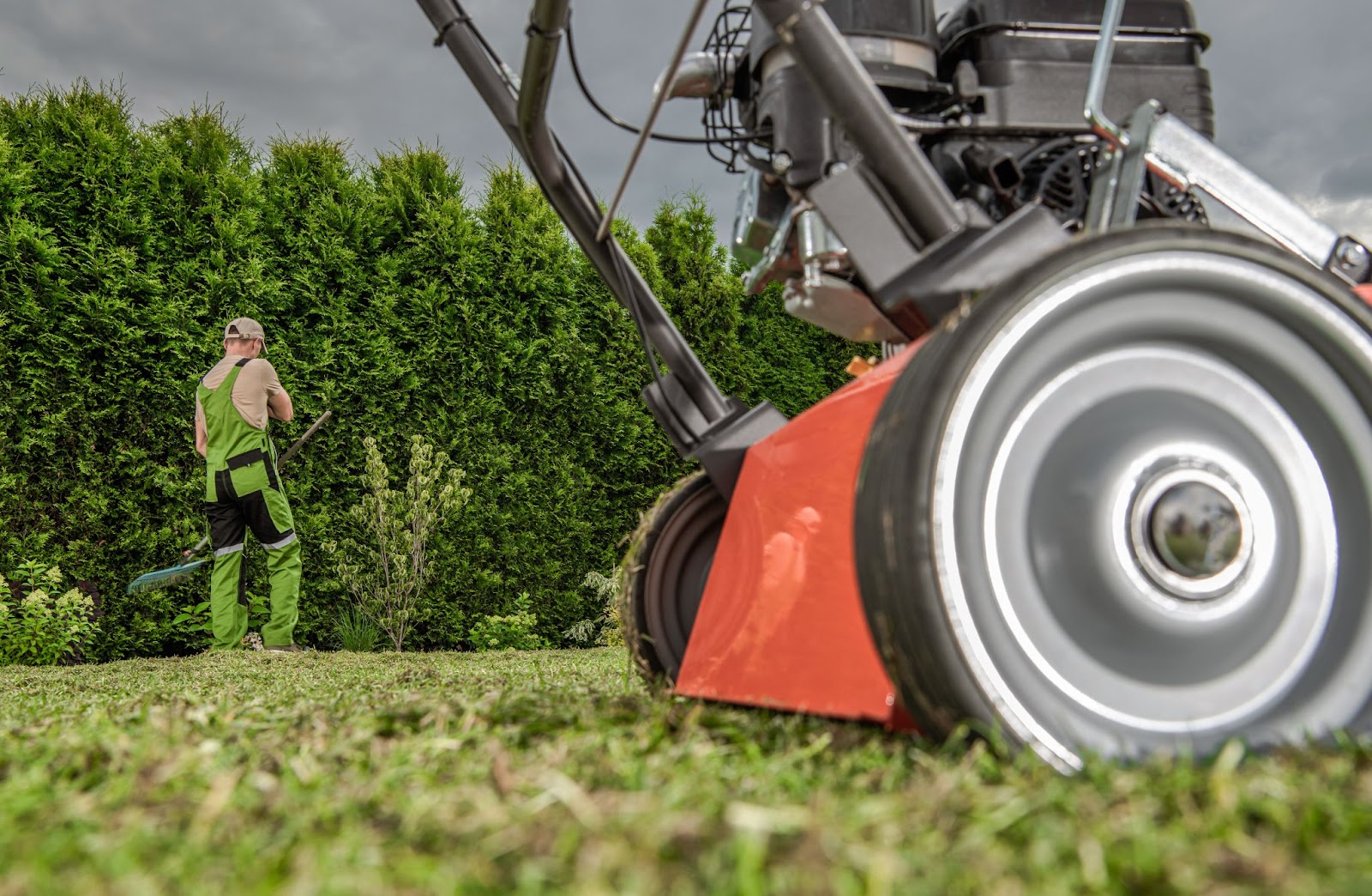 Lawn aerator equipment in foreground with a man trimming hedges in the background, showing tools and tasks included in advanced summer lawn treatment services.